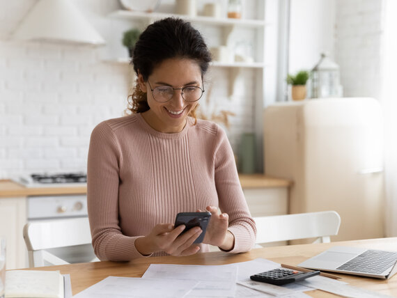 Smiling young Caucasian woman manage household budget finances paying bills taxes on smartphone online. Happy female busy with documents calculate expenses expenditures on cellphone on internet.